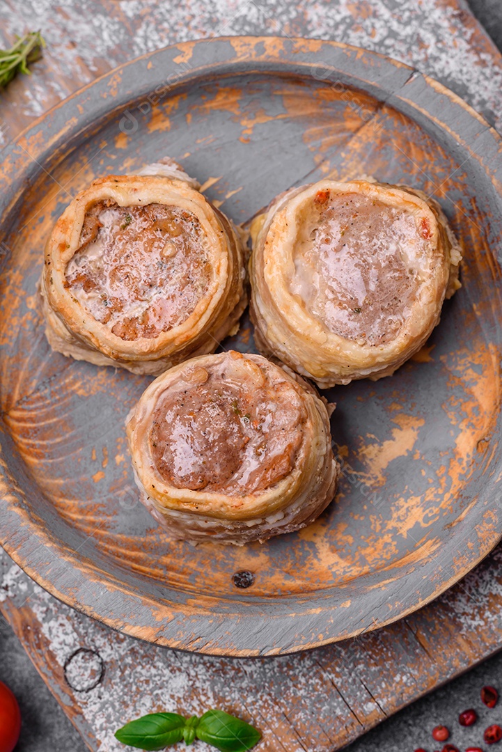 Deliciosos rolos de carne de porco ou frango crus e frescos embrulhados em bacon, com sal, especiarias e ervas sobre um fundo escuro de concreto