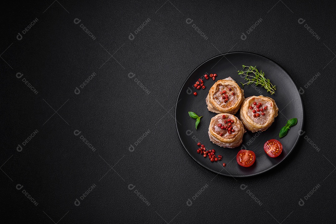 Deliciosos rolos de carne de porco ou frango crus e frescos embrulhados em bacon, com sal, especiarias e ervas sobre um fundo escuro de concreto