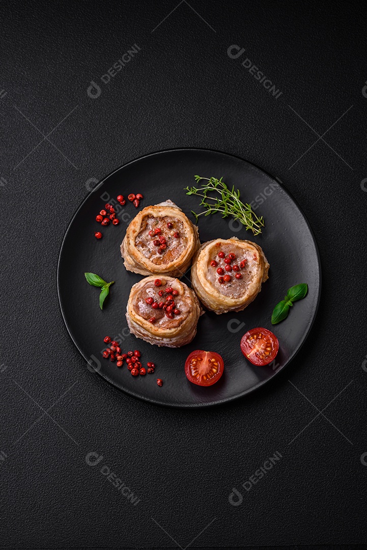 Deliciosos rolos de carne de porco ou frango crus e frescos embrulhados em bacon, com sal, especiarias e ervas sobre um fundo escuro de concreto