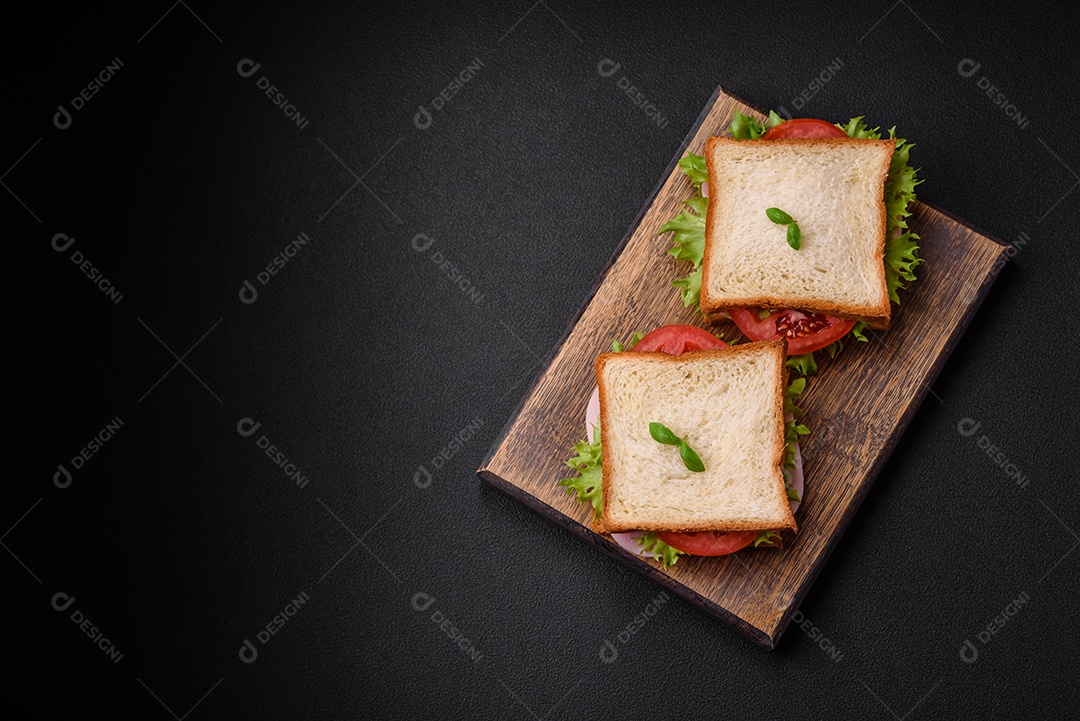 Delicioso sanduíche com torradas, presunto, tomate, queijo e alface com sal, especiarias e ervas sobre fundo escuro de concreto