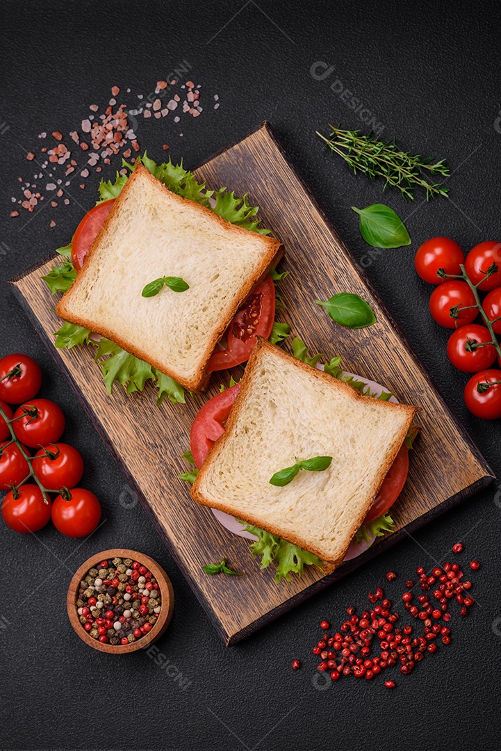 Delicioso sanduíche com torradas, presunto, tomate, queijo e alface com sal, especiarias e ervas sobre fundo escuro de concreto