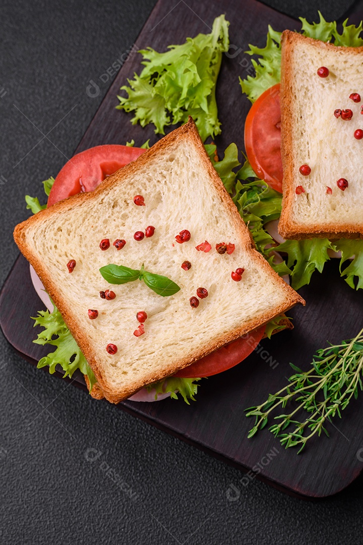 Delicioso sanduíche com torradas, presunto, tomate, queijo e alface com sal, especiarias e ervas sobre fundo escuro de concreto