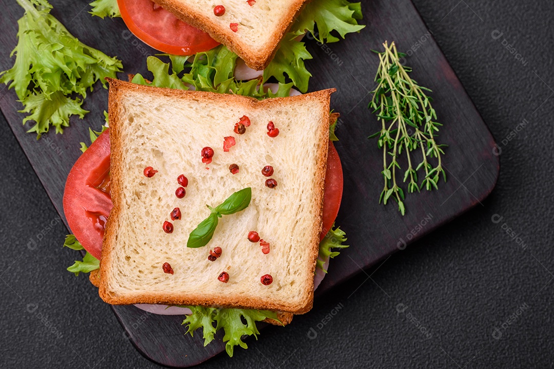 Delicioso sanduíche com torradas, presunto, tomate, queijo e alface com sal, especiarias e ervas sobre fundo escuro de concreto