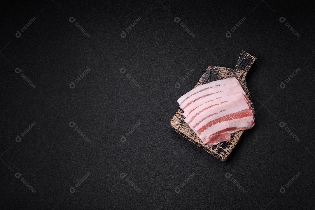 Bacon cru fresco cortado em fatias com sal, especiarias e ervas sobre um fundo de concreto texturizado escuro