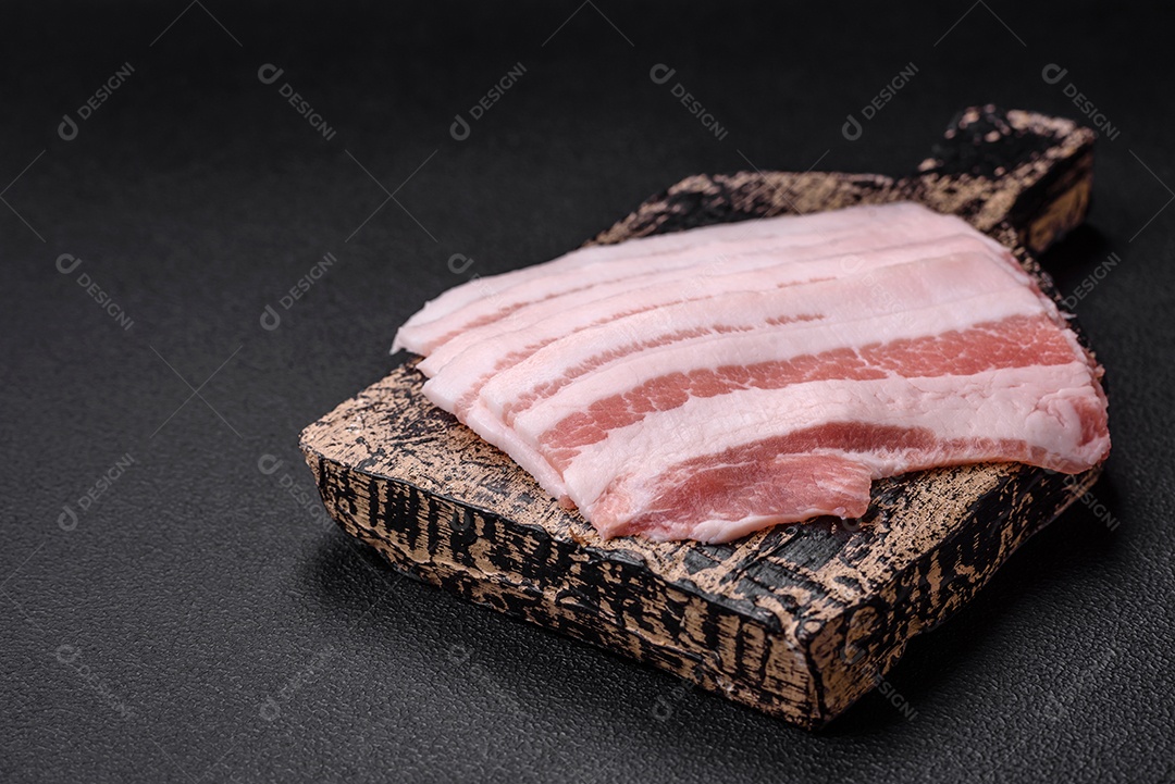Bacon cru fresco cortado em fatias com sal, especiarias e ervas sobre um fundo de concreto texturizado escuro