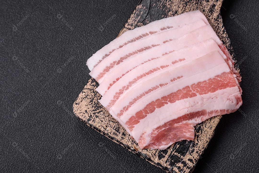 Bacon cru fresco cortado em fatias com sal, especiarias e ervas sobre um fundo de concreto texturizado escuro