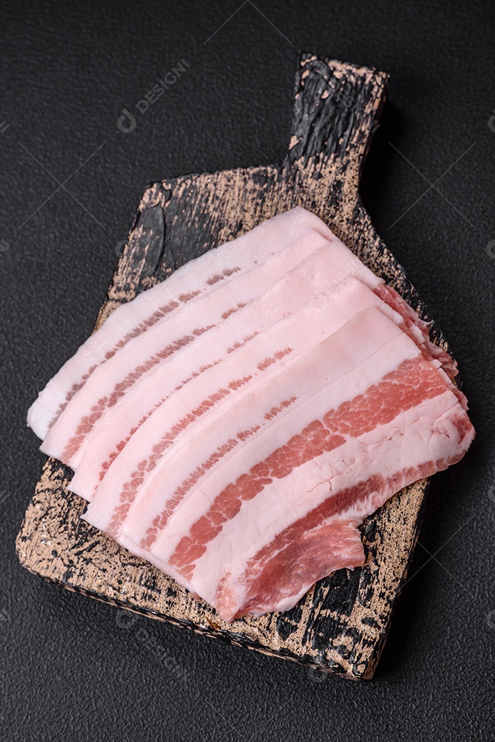 Bacon cru fresco cortado em fatias com sal, especiarias e ervas sobre um fundo de concreto texturizado escuro
