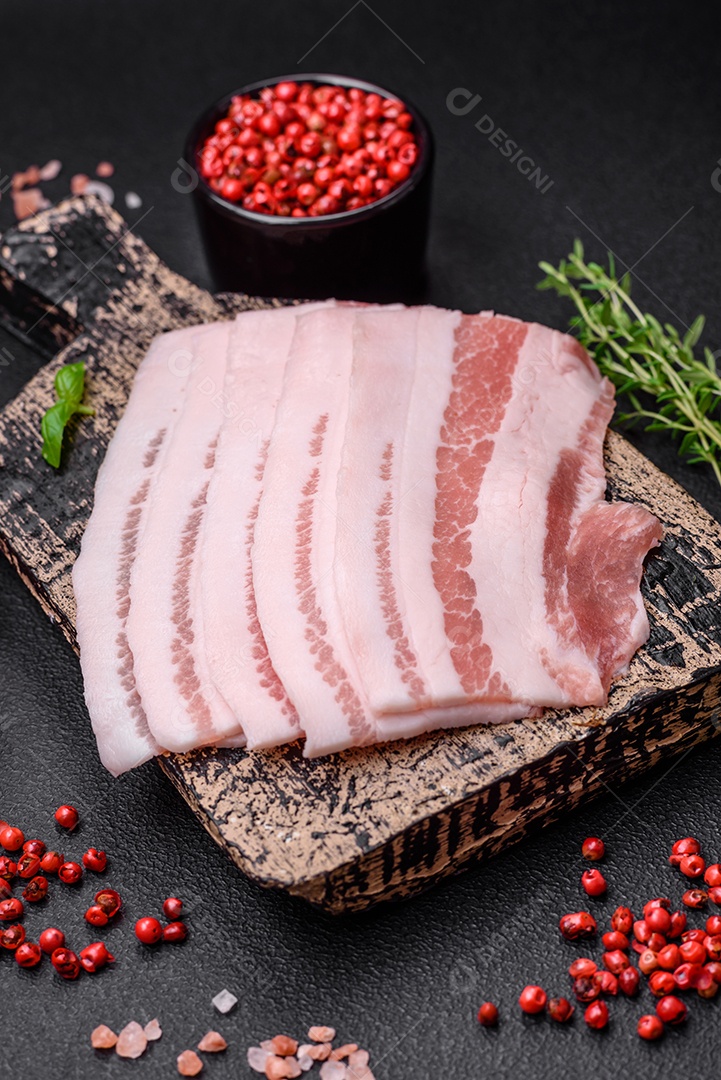 Bacon cru fresco cortado em fatias com sal, especiarias e ervas sobre um fundo de concreto texturizado escuro