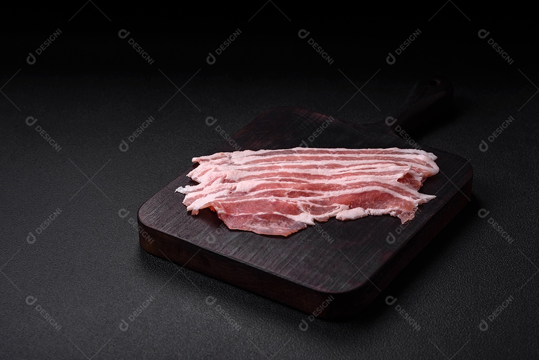 Bacon cru fresco cortado em fatias com sal, especiarias e ervas sobre um fundo de concreto texturizado escuro