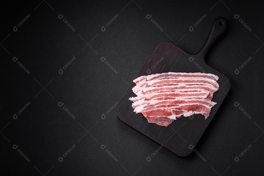 Bacon cru fresco cortado em fatias com sal, especiarias e ervas sobre um fundo de concreto texturizado escuro
