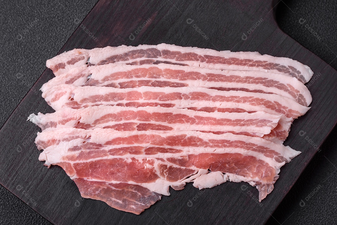 Bacon cru fresco cortado em fatias com sal, especiarias e ervas sobre um fundo de concreto texturizado escuro