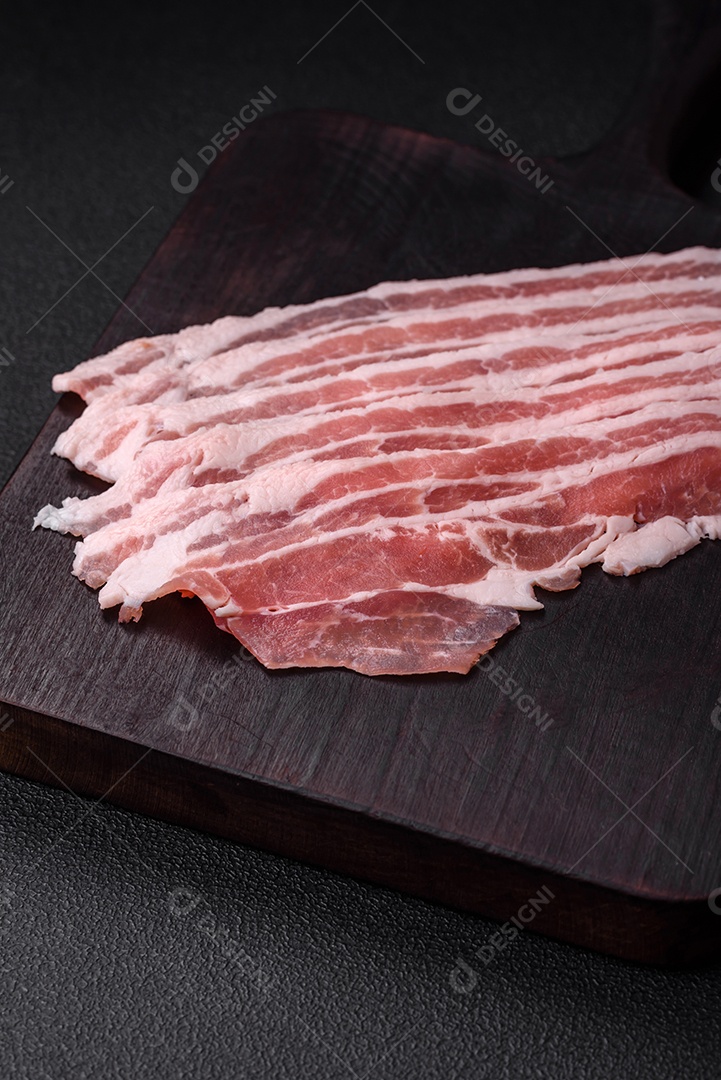 Bacon cru fresco cortado em fatias com sal, especiarias e ervas sobre um fundo de concreto texturizado escuro