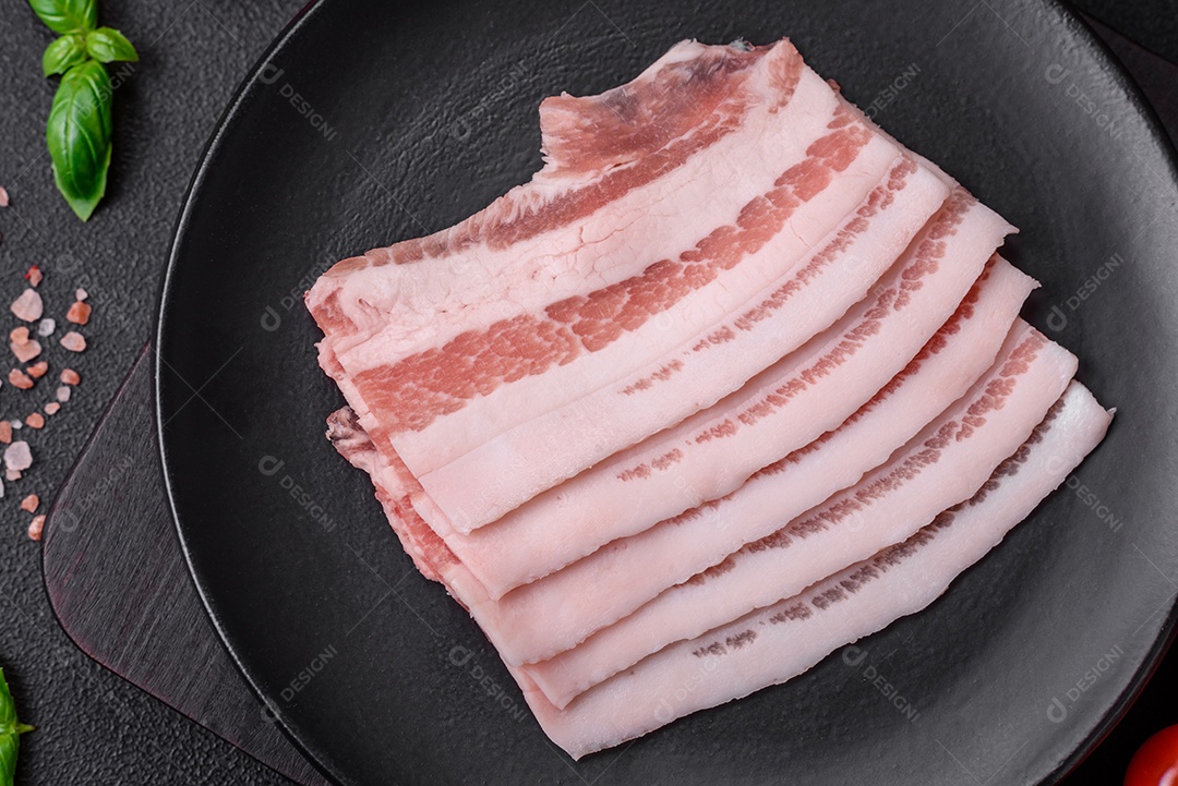 Bacon cru fresco cortado em fatias com sal, especiarias e ervas sobre um fundo de concreto texturizado escuro