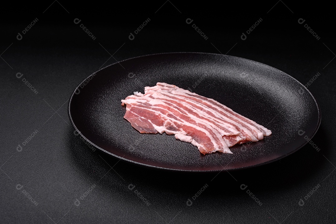 Bacon cru fresco cortado em fatias com sal, especiarias e ervas sobre um fundo de concreto texturizado escuro