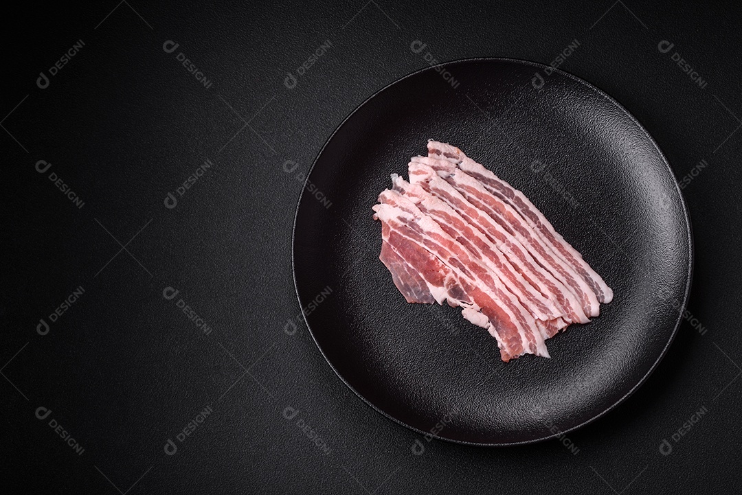 Bacon cru fresco cortado em fatias com sal, especiarias e ervas sobre um fundo de concreto texturizado escuro