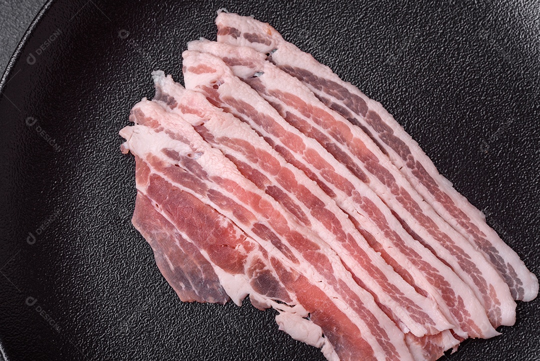 Bacon cru fresco cortado em fatias com sal, especiarias e ervas sobre um fundo de concreto texturizado escuro