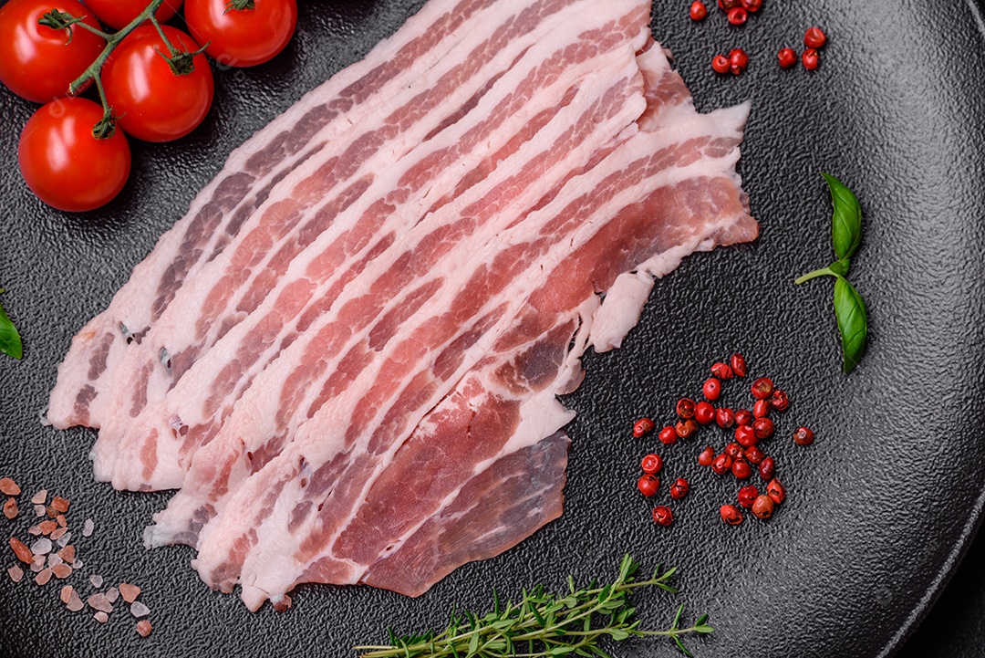 Bacon cru fresco cortado em fatias com sal, especiarias e ervas sobre um fundo de concreto texturizado escuro