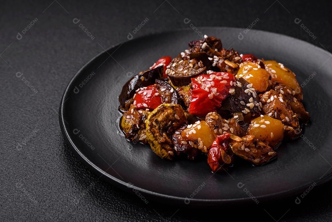Deliciosa salada fresca e suculenta de berinjela assada, tomate, pimentão, sementes de gergelim, especiarias e sal sobre um fundo escuro de concreto