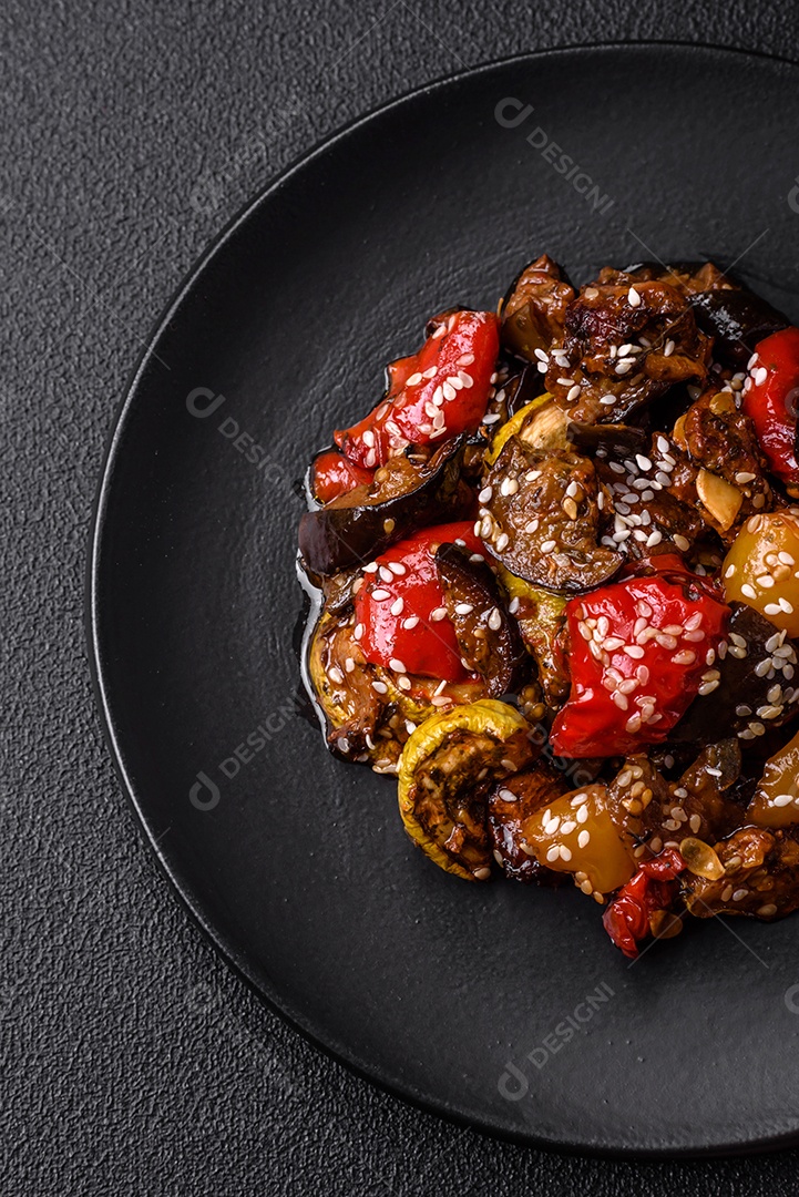 Deliciosa salada fresca e suculenta de berinjela assada, tomate, pimentão, sementes de gergelim, especiarias e sal sobre um fundo escuro de concreto