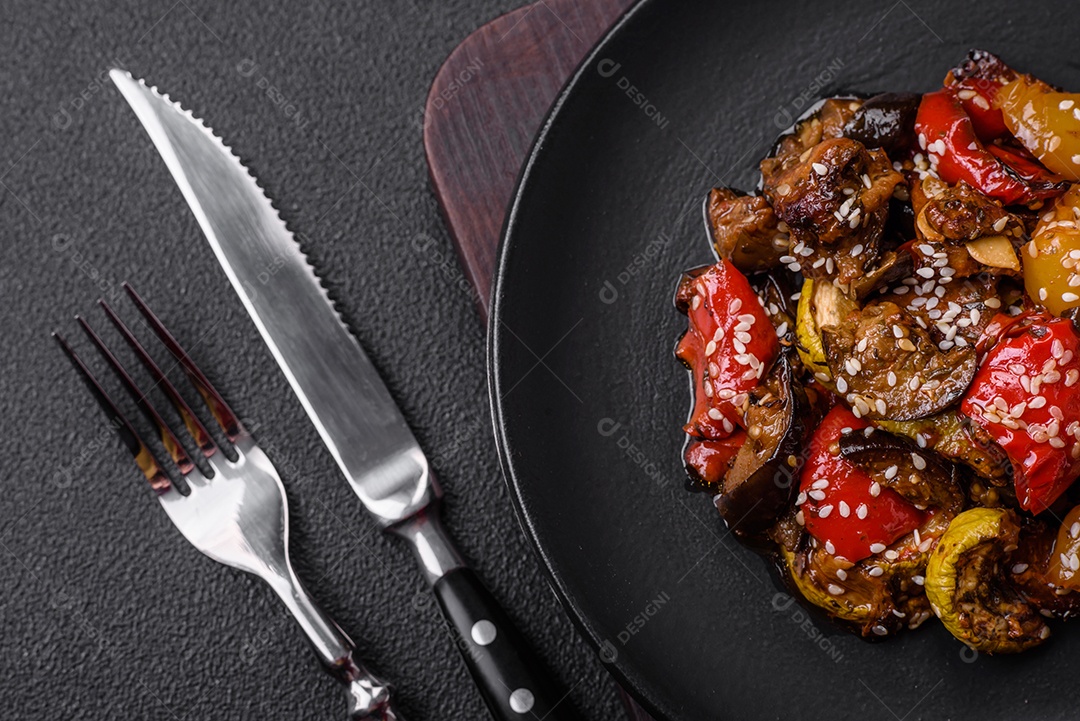 Deliciosa salada fresca e suculenta de berinjela assada, tomate, pimentão, sementes de gergelim, especiarias e sal sobre um fundo escuro de concreto