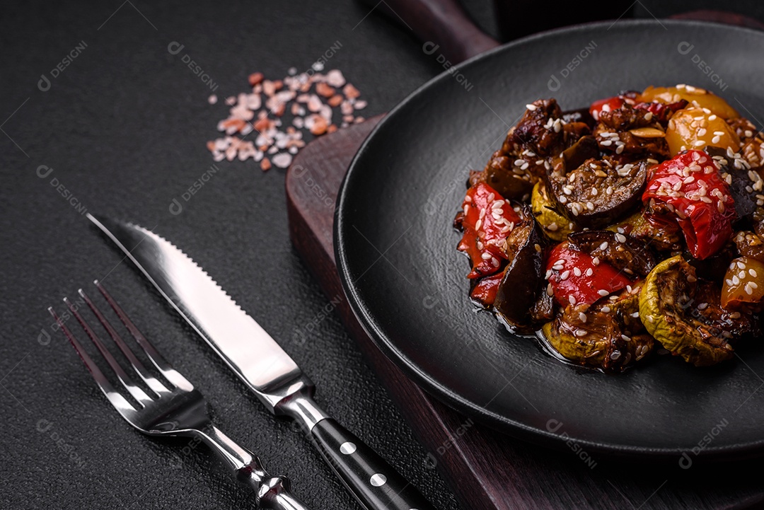 Deliciosa salada fresca e suculenta de berinjela assada, tomate, pimentão, sementes de gergelim, especiarias e sal sobre um fundo escuro de concreto