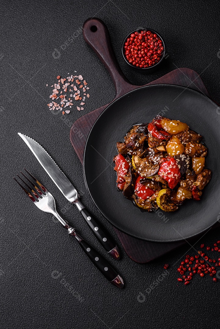 Deliciosa salada fresca e suculenta de berinjela assada, tomate, pimentão, sementes de gergelim, especiarias e sal sobre um fundo escuro de concreto