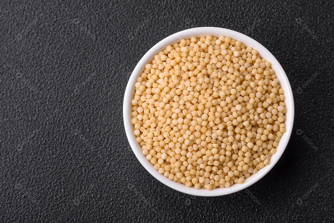 Massa crua ptitim em uma tigela de cerâmica sobre um fundo escuro de concreto. Preparação de comida israelense