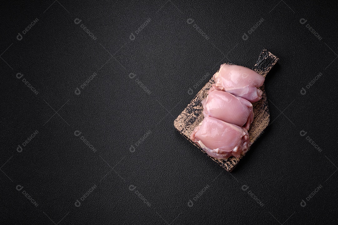 Filetes de coxa de frango cru fresco com sal, especiarias e ervas sobre um fundo escuro de concreto