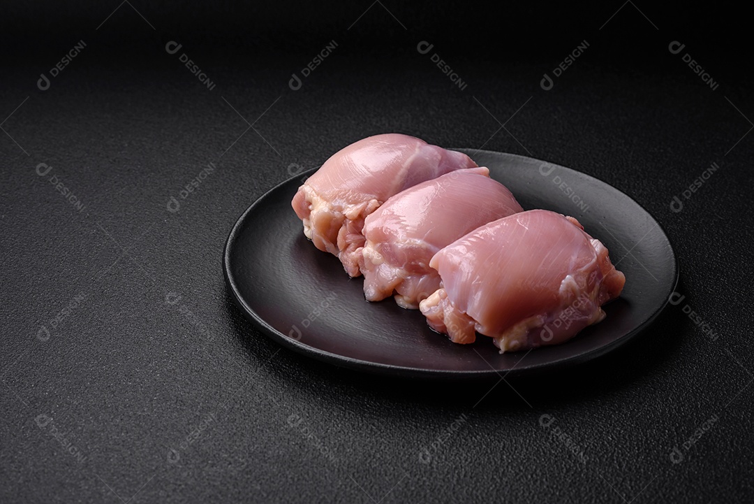 Filetes de coxa de frango cru fresco com sal, especiarias e ervas sobre um fundo escuro de concreto