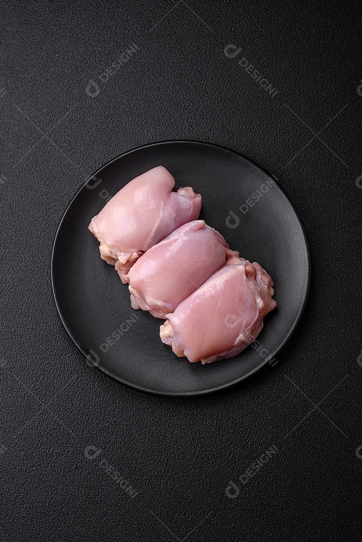 Filetes de coxa de frango cru fresco com sal, especiarias e ervas sobre um fundo escuro de concreto