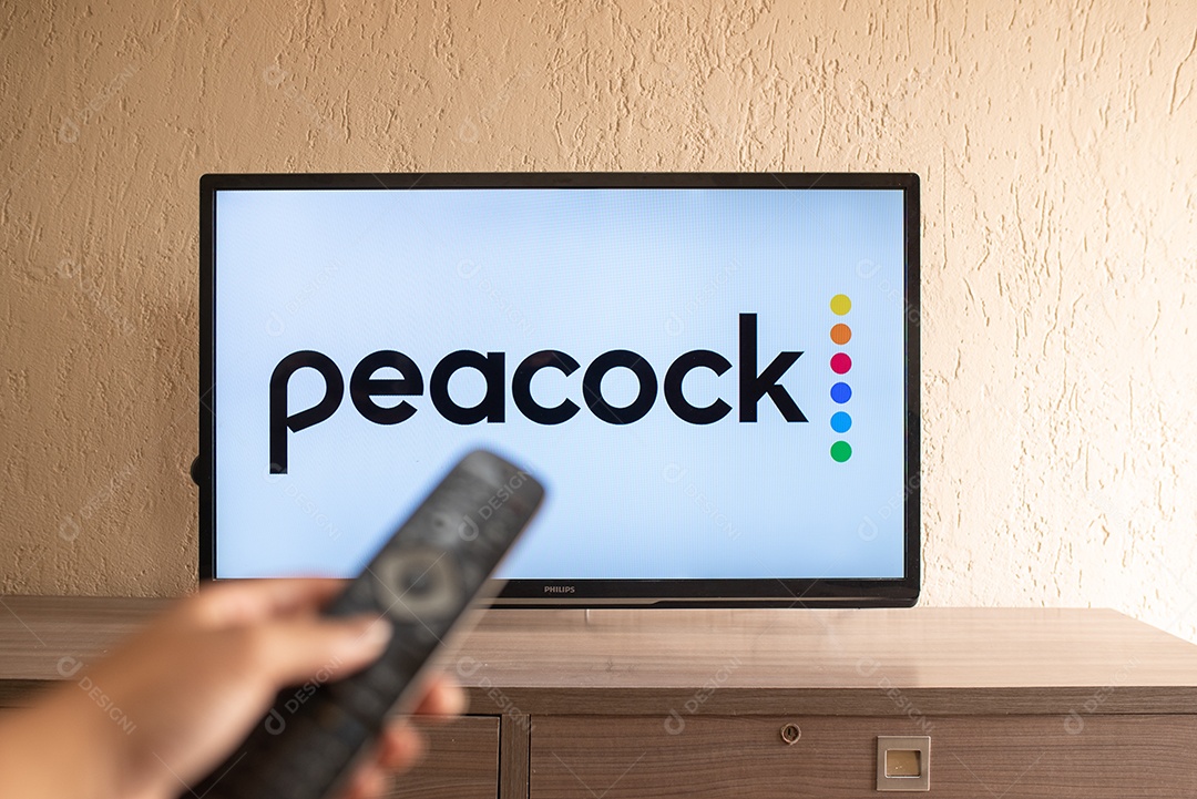 Mão segurando um controle remoto de TV visto na frente do logotipo do Peacock