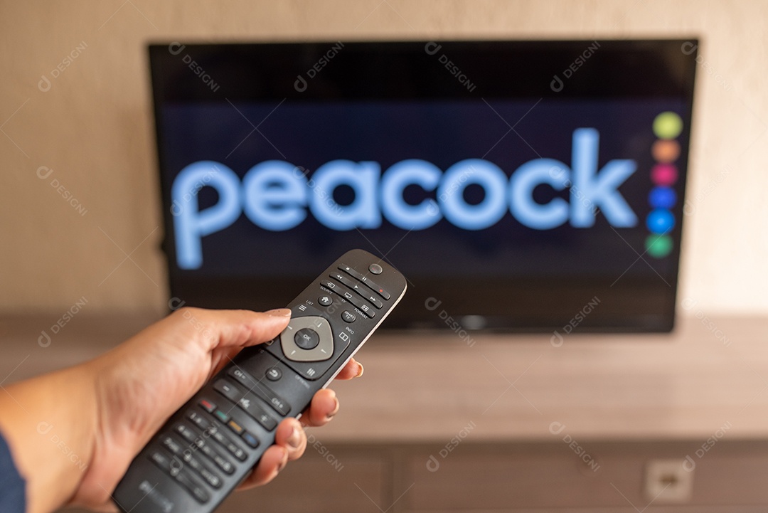Mão segurando um controle remoto de TV visto na frente do logotipo do Peacock