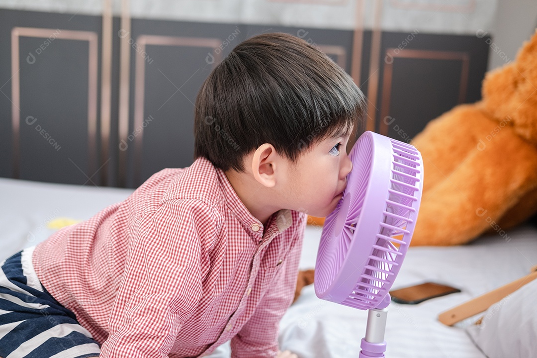 Menino asiático deitado em um dia quente brincando alegremente com um ventilador portátil