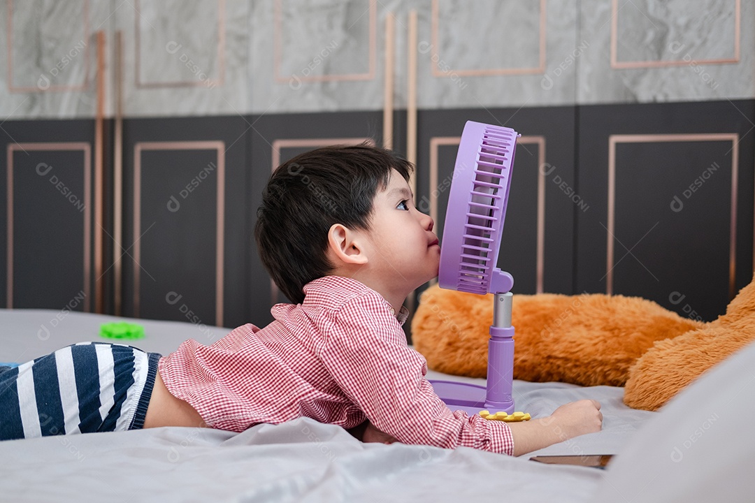 Menino asiático deitado em um dia quente brincando alegremente com um ventilador portátil