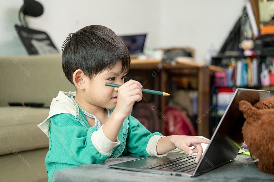 Um garotinho asiático assistindo aula para fazer tarefa no laptop