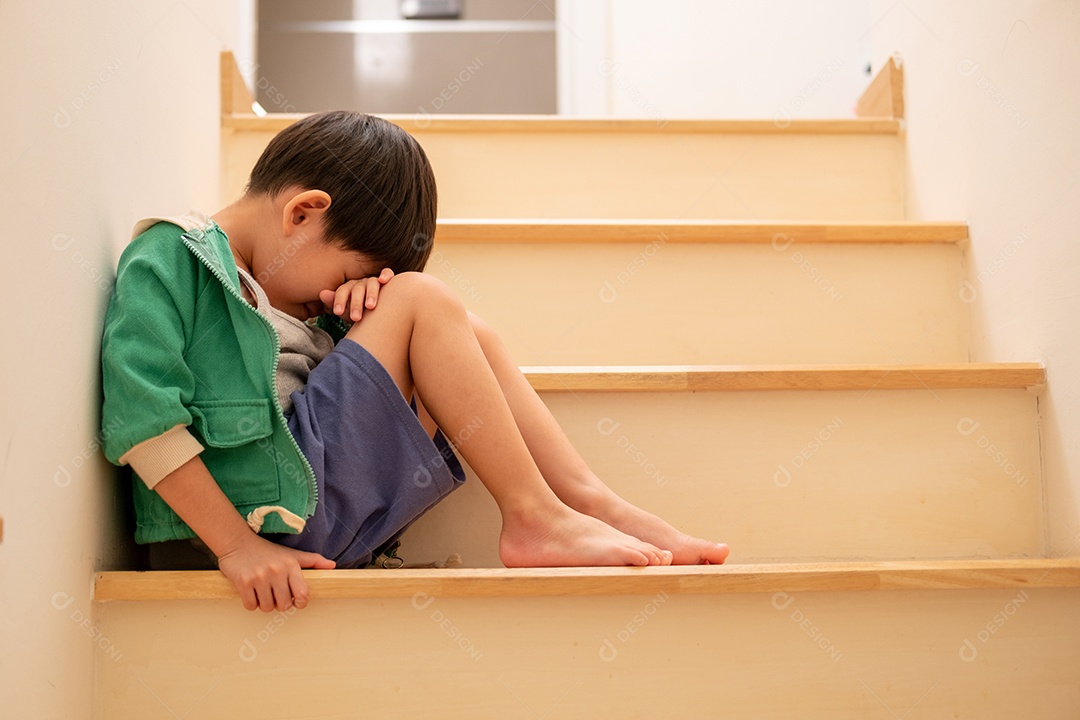 Um menino trsite asiático está sentado nos degraus com cabeça a baixa