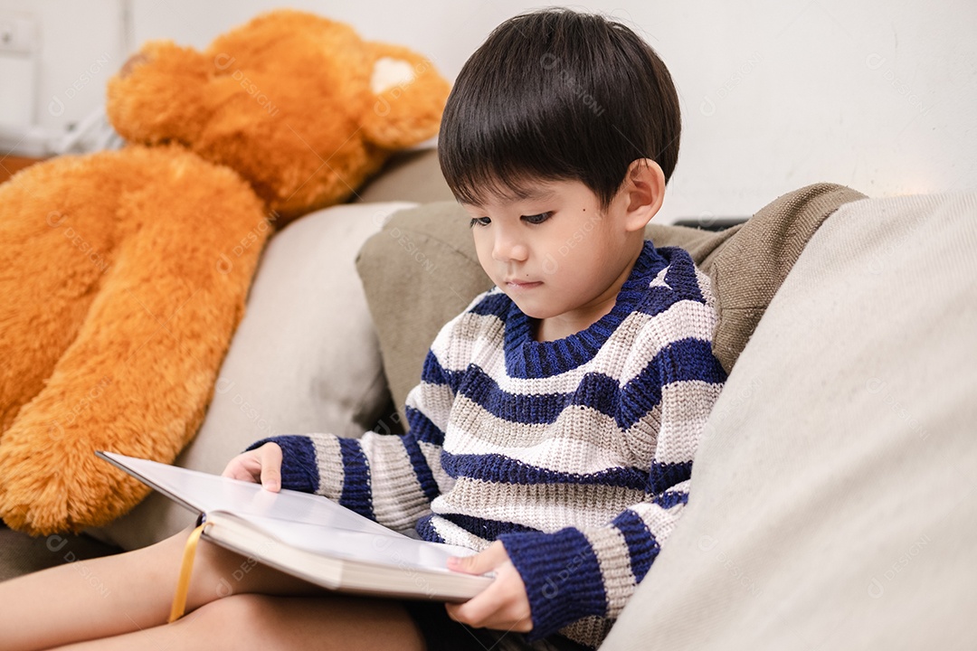 Menino asiático lendo um livro no sofá aprendendo fora da sala de aula