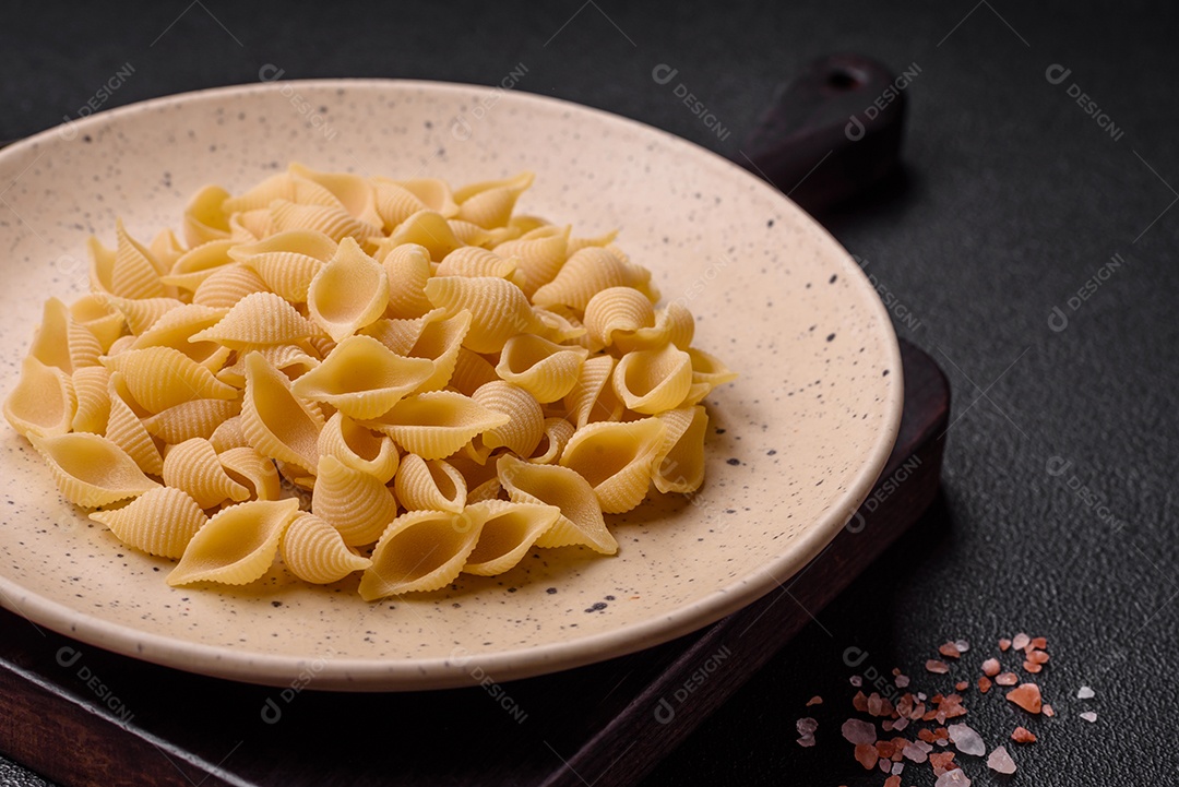 Conchiglie de massa italiana crua de trigo duro com legumes, sal e especiarias sobre um fundo escuro de concreto