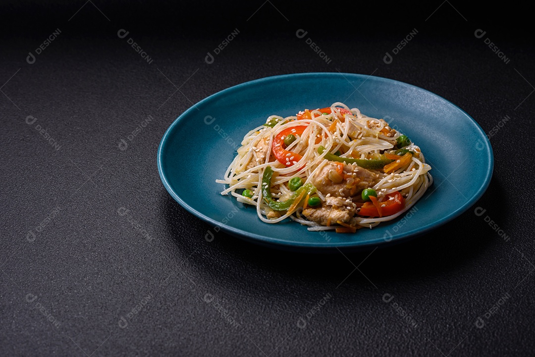 Saboroso prato de culinária asiática com macarrão de arroz, frango, aspargos, pimenta, sementes de gergelim e molho de soja em fundo escuro de concreto