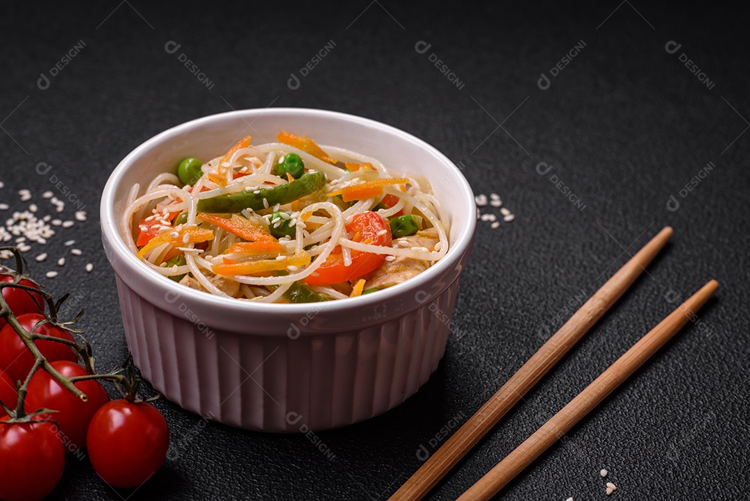 Saboroso prato de culinária asiática com macarrão de arroz, frango, aspargos, pimenta, sementes de gergelim e molho de soja em fundo escuro de concreto