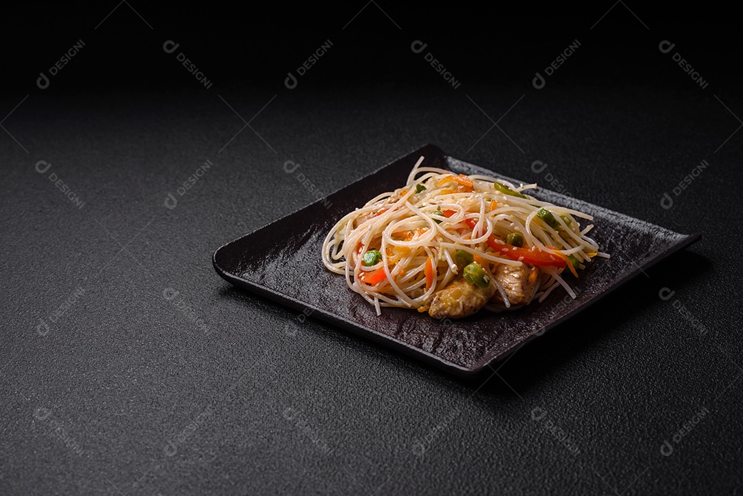 Saboroso prato de culinária asiática com macarrão de arroz, frango, aspargos, pimenta, sementes de gergelim e molho de soja em fundo escuro de concreto