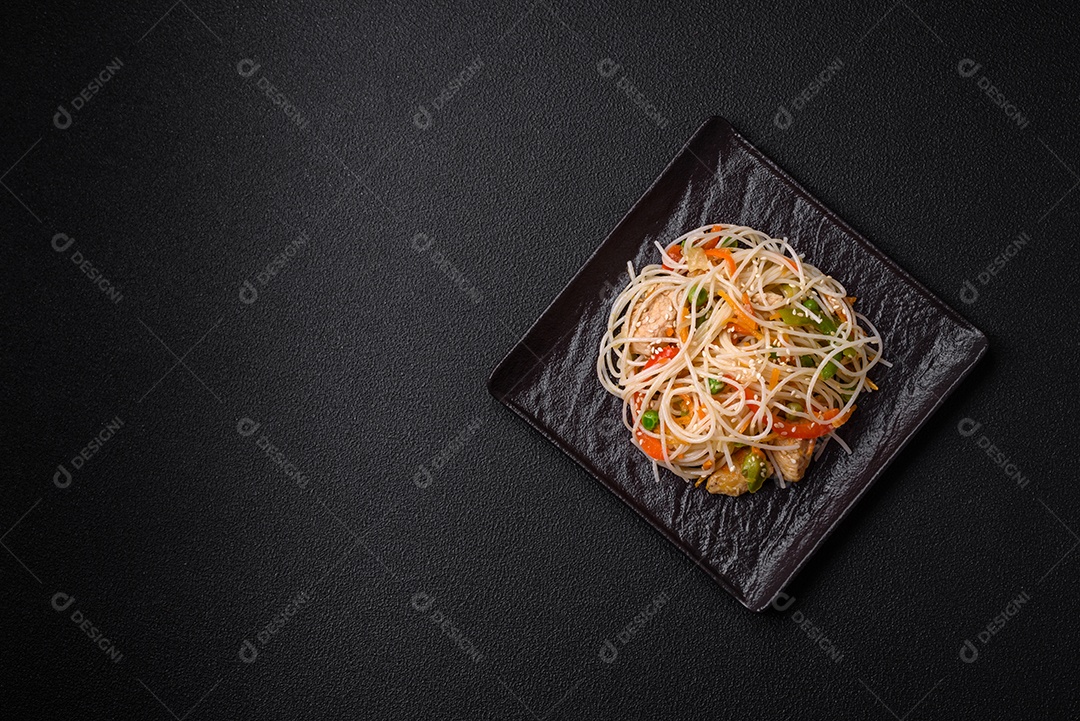 Saboroso prato de culinária asiática com macarrão de arroz, frango, aspargos, pimenta, sementes de gergelim e molho de soja em fundo escuro de concreto