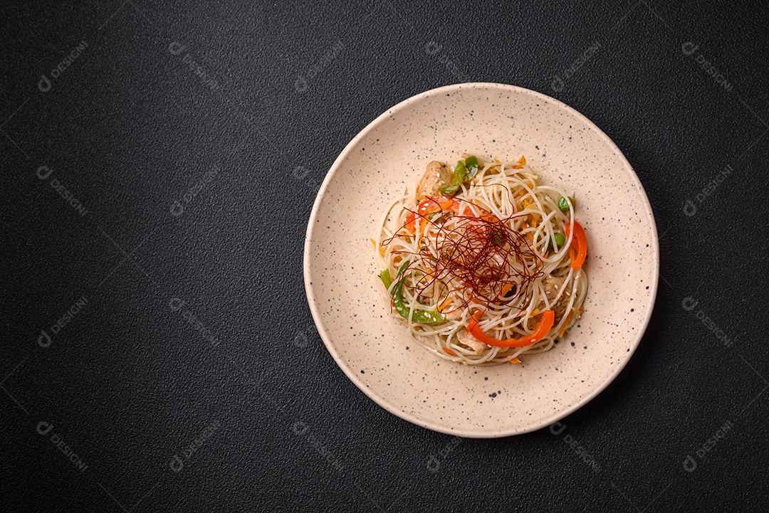 Saboroso prato de culinária asiática com macarrão de arroz, frango, aspargos, pimenta, sementes de gergelim e molho de soja em fundo escuro de concreto