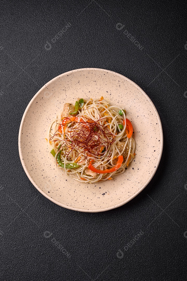 Saboroso prato de culinária asiática com macarrão de arroz, frango, aspargos, pimenta, sementes de gergelim e molho de soja em fundo escuro de concreto