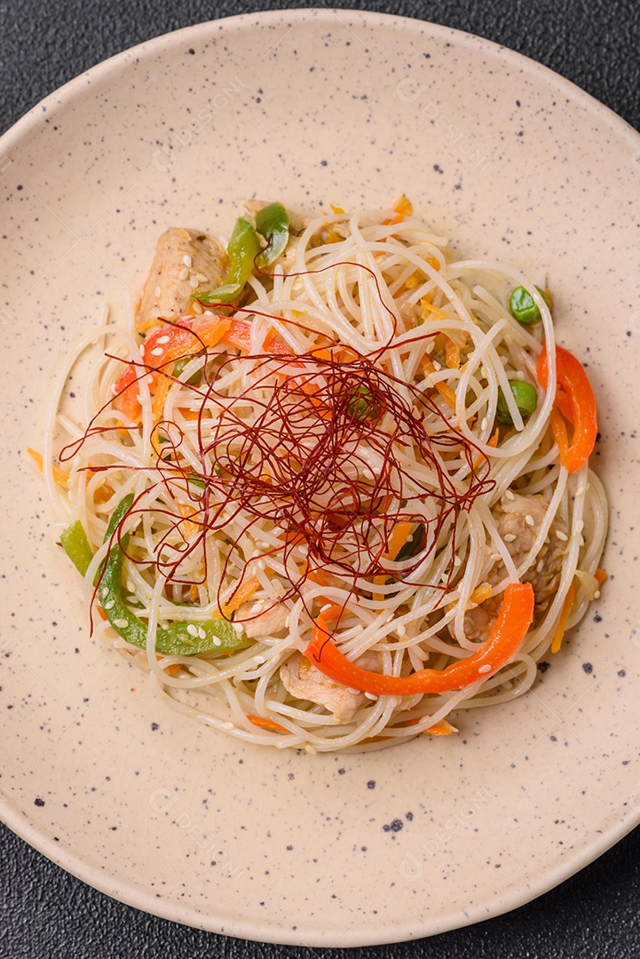 Saboroso prato de culinária asiática com macarrão de arroz, frango, aspargos, pimenta, sementes de gergelim e molho de soja em fundo escuro de concreto