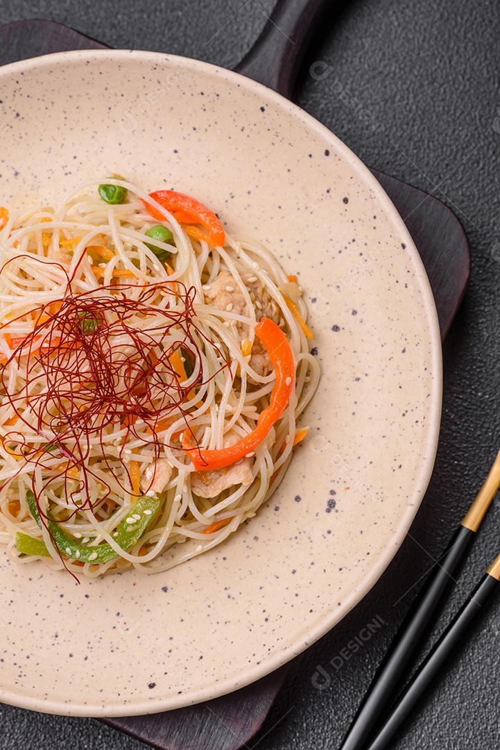 Saboroso prato de culinária asiática com macarrão de arroz, frango, aspargos, pimenta, sementes de gergelim e molho de soja em fundo escuro de concreto