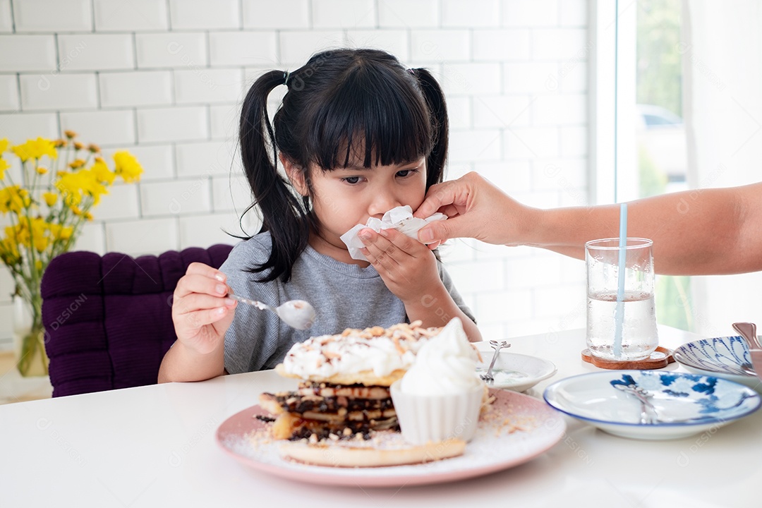 A mão da mãe está usando um lenço de papel para limpar a boca da filha asiática de manchas de comida enquanto come à mesa.