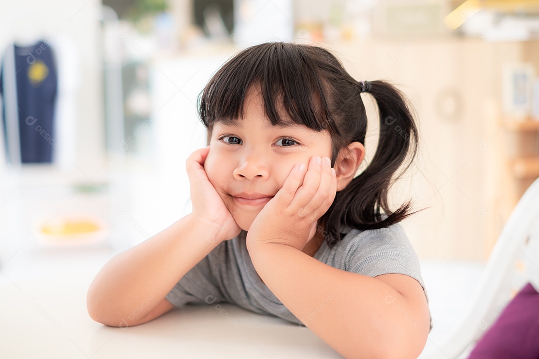 Linda menina asiática sentada com a mão no queixo e sorrindo enquanto espera para comer em uma mesa em um restaurante.
