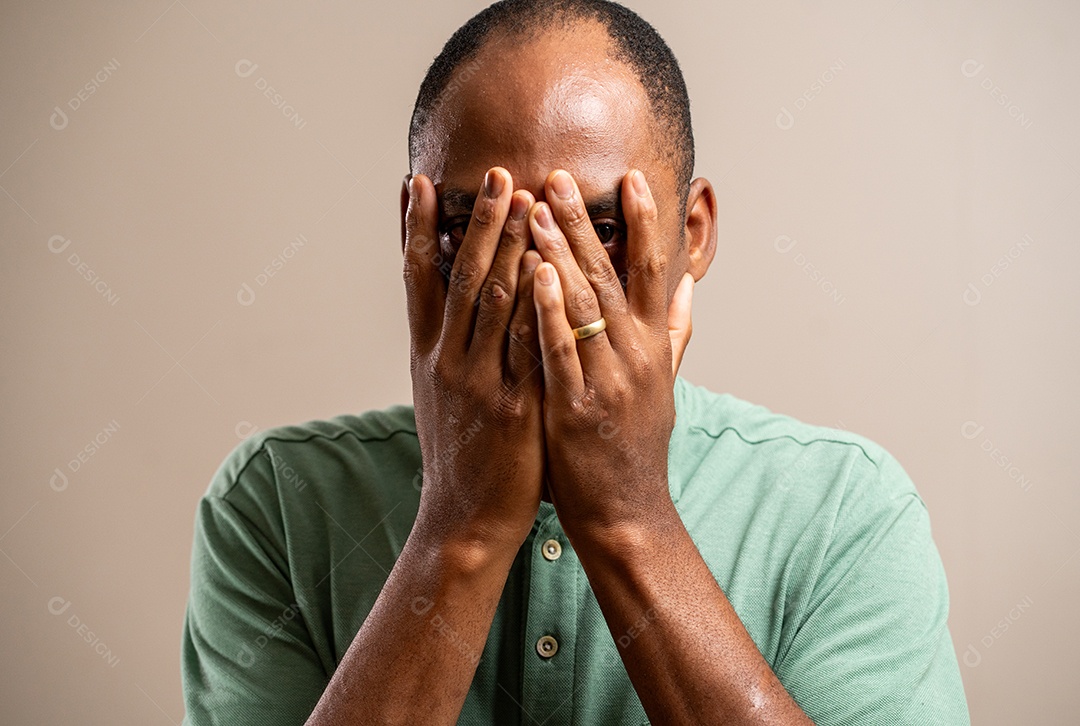 Homem com as mãos no rosto, olhando entre os dedos, sobre fundo pastel.