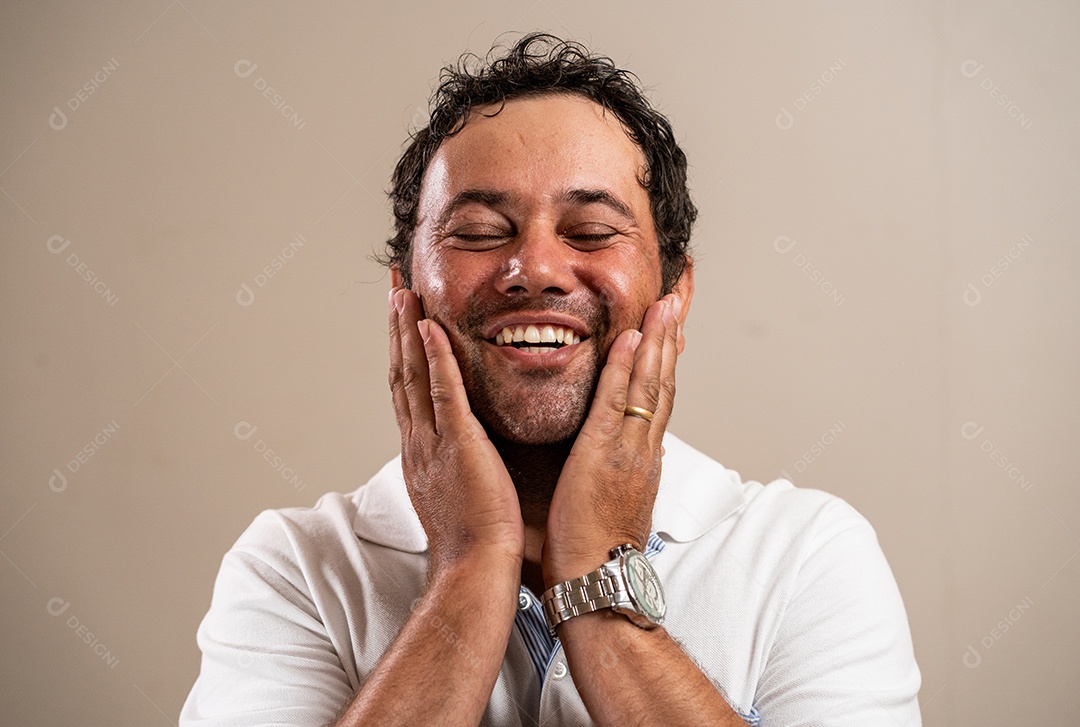 Homem com barba por fazer sorrindo com a mão no rosto e olhos fechados em fundo pastel.